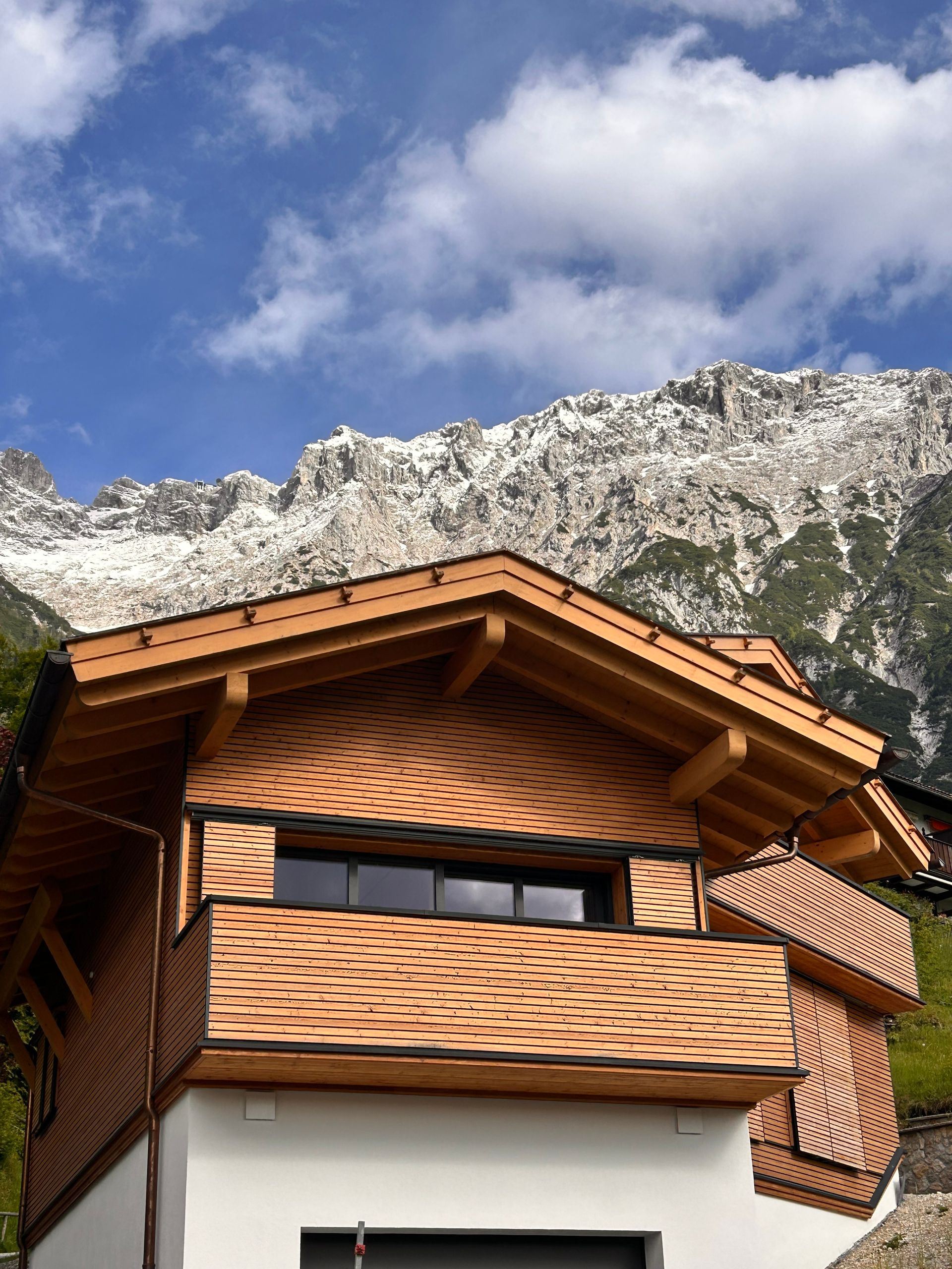 A newly built alpine chalet with horizontal timber cladding and deep eaves, below a snow-dusted rock face